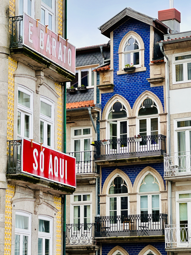 Colorful buildings in Porto, Portugal. One is covered in blue tile. Another is covered in yellow tile with a sign that says So Aqui.
