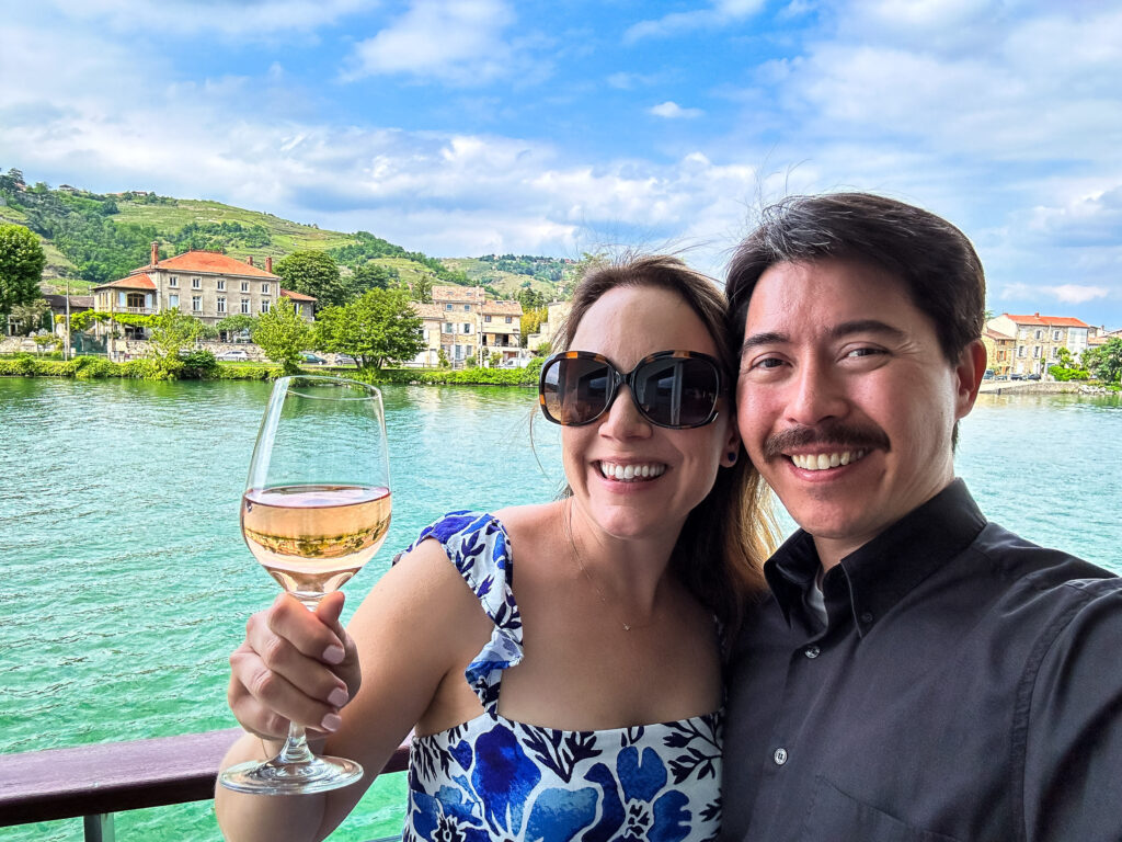 Rachelle and Pete on their cruise balcony on the Rhone. Rachelle is holding a glass of rose wine and there's a gorgeous provincial house on the coastline.