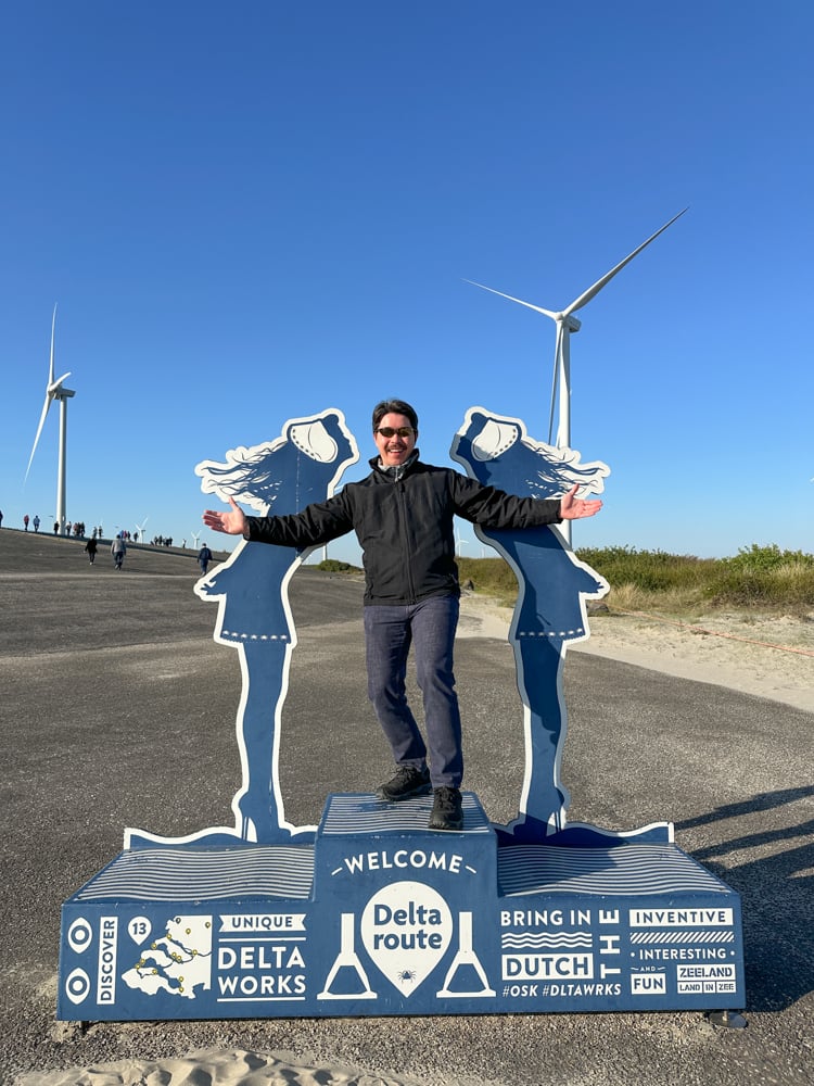 Pete standing next to sign of the Delta Route with large modern windmills behind him during the Tulips and Windmills river cruise