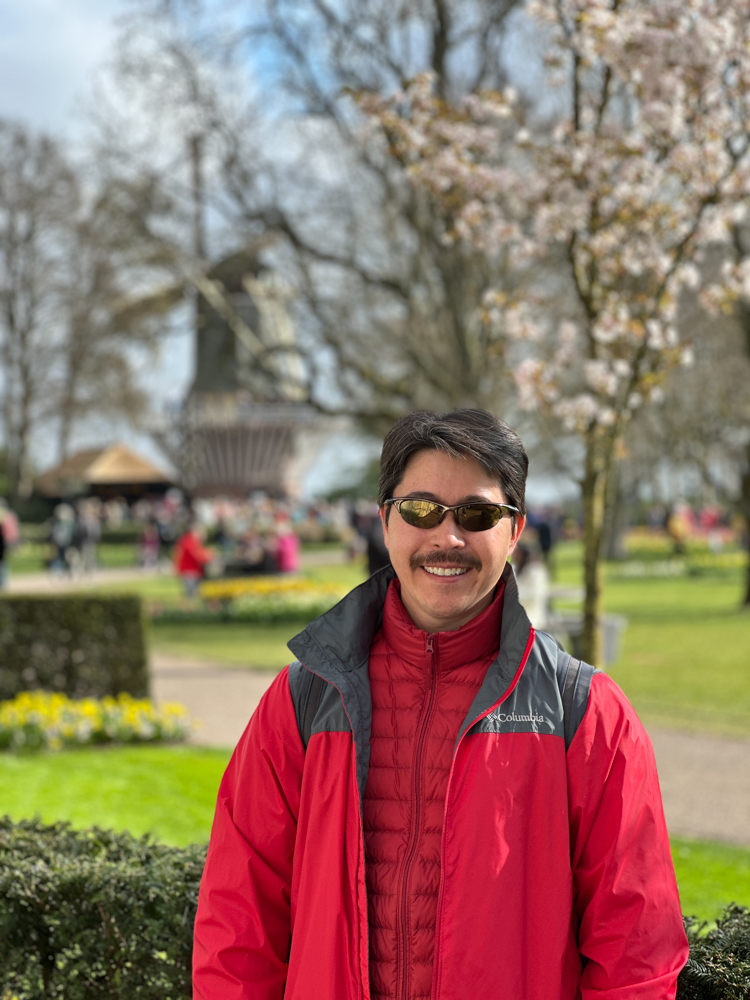 Pete in a red jacket standing under cherry blossoms with a windmill in the background at Keukenhof Gardens