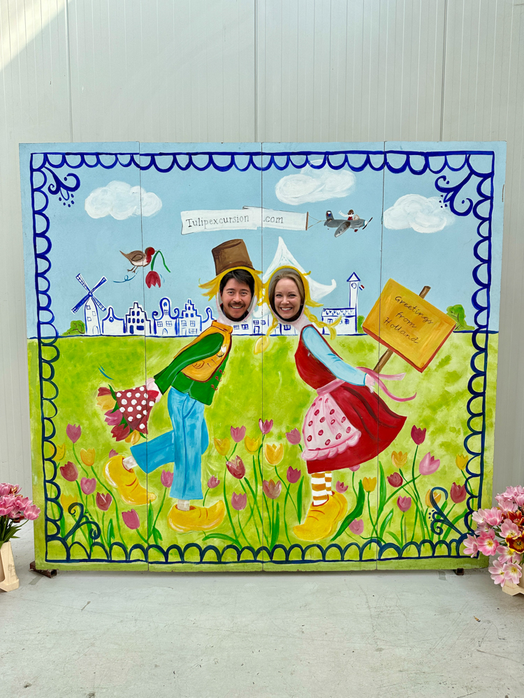 Pete and Rachelle posing behind a mural of a Dutch countryside with their heads in holes to put their faces in on the cartoon bodies of the farmers in the mural.